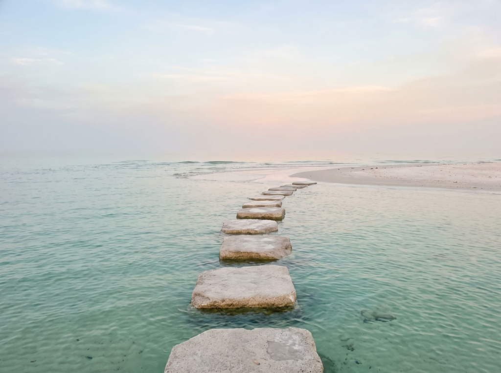 Flat stepping stones crossing calm, clear shallow water toward a sandy shore