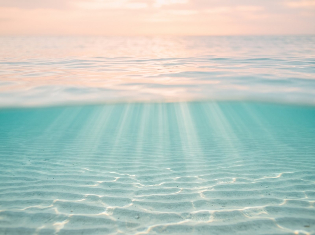 Shafts of sunlight reach through shallow turquoise water, lighting rippled pale sand below a calm surface.